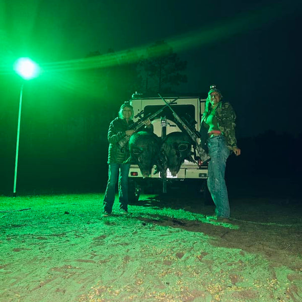 Load image into Gallery viewer, Two hunters standing next to a truck with hogs under a green light after night huning.